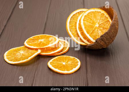 Getrocknete Orangenscheiben sind auf dem Tisch verstreut. Orangenchips in einer Kokosnussschale. Dunkler Holzhintergrund. Stockfoto