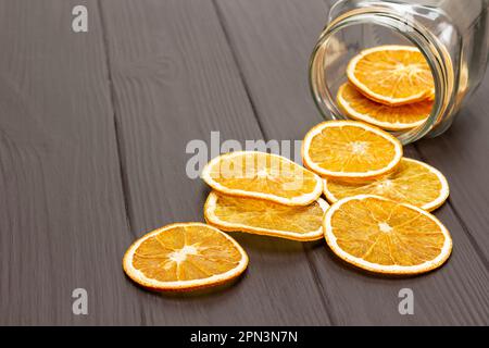 Getrocknete Orangenscheiben werden aus einem Glasgefäß auf dem Tisch gestreut. Speicherplatz kopieren. Dunkler Holzhintergrund. Stockfoto