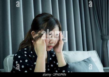 Aufnahme einer jungen Frau, die ihren Kopf aufgrund von Schmerzen in Unbehagen hält, während sie Migräne oder schreckliche Kopfschmerzen fühlt, Schmerzen erleidet, die Stirnrunzeln der Massagebügel massiert Stockfoto
