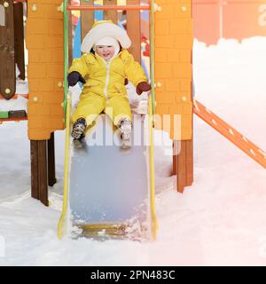 Der kleine Junge fährt auf einer Rutsche und spielt auf einem Winterspielplatz. Ein Kind in einem gelben Overall auf einer Kinderrutsche im Schnee. Ein Kind im Alter von einem Jahr Stockfoto