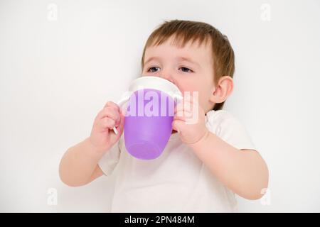 Fröhliches Baby trinkt Wasser aus einer Tasse auf weißem Studiohintergrund. Ruhendes Kind mit Tassensaft in den Händen. Kind etwa zwei Jahre alt (ein Jahr neun Monate) Stockfoto