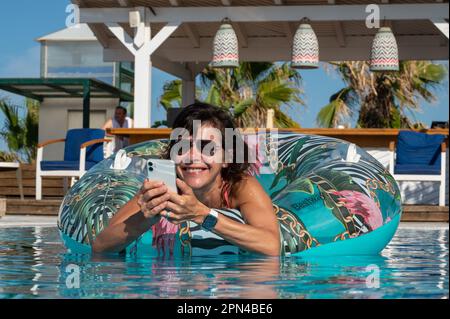 Aktive 60-jährige Frau, die ein Smartphone benutzt und im Urlaub im Pool lebt Stockfoto