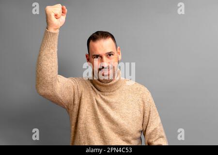 Bärtiger hispanischer Mann in seinen 40s Jahren mit beigefarbenem Rollkragenpullover, der seine Faust kräftig erhebt und einen großen Sieg feiert, isoliert vor grauem Hintergrund Stockfoto