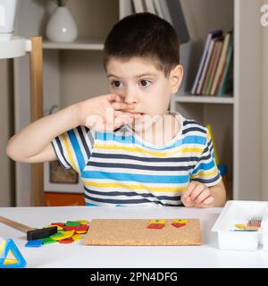 Ein 5-jähriger Junge beißt sich die Nägel, während er an einem Tisch sitzt und im Kinderzimmer mit Mosaiken spielt. Schlechte Angewohnheiten bei Kindern Stockfoto