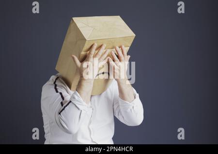 Ein trauriger Mann in einem weißen Hemd mit einer Tasche auf dem Kopf, mit einem gezeichneten traurigen Emoticon, hat Angst und schließt seine Augen mit seinen Händen. Emotionen und Gesten. Stockfoto