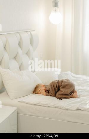 Ein Junge im braunen Bademantel, liegt auf einem weißen Bett, bedeckt sein Gesicht mit den Händen. Stockfoto