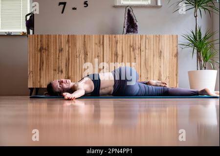Junge Frau, die nach einem Heimtraining auf dem Rücken liegt. Dehnen, während sie ihren Körper verdreht, um die Muskeln zu entspannen. In einem Wohnzimmer. Stockfoto