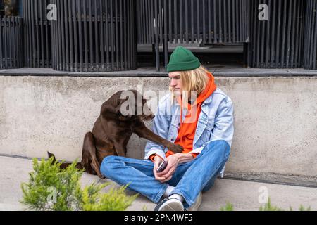 Ein freundlicher, obdachloser Hund, der mit einem Kerl in der Innenstadt sitzt und um Essen bettelt und ihm die Pfote gibt. Stockfoto