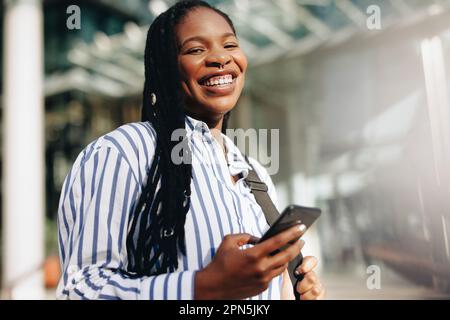 Porträt einer jungen schwarzen Geschäftsfrau, die auf dem Weg zur Arbeit in der Stadt vor der Kamera lächelt. Glückliche junge Geschäftsfrau, die während der Kommunikation ein Smartphone benutzt Stockfoto