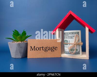 Spielzeughaus, grüne Pflanze und Banknoten mit dem Wort Hypothek auf blauem Hintergrund. Stockfoto