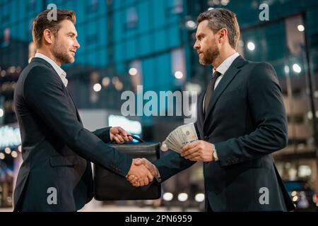 Wir zählen uns Dollarscheine, Geld, Finanzen, Sparen, Bankwesen und Mitarbeiterkonzept. Ein Geschäftsmann, der einem Partner im Hintergrund einer Nachtstadt Geld spendet. Zwei Stockfoto