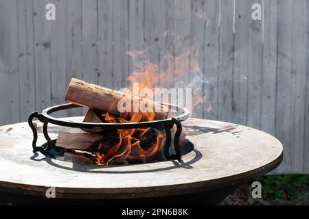 Runder Grill in Form einer Schüssel mit Feuer im Inneren. Ein Mann bereitet ein Herd für ein Barbecue vor. Brennholz brennt in der Schüssel des Herds. Stockfoto