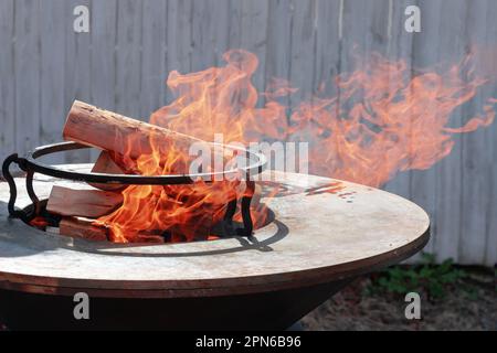 Runder Grill in Form einer Schüssel mit Feuer im Inneren. Ein Mann bereitet ein Herd für ein Barbecue vor. Brennholz brennt in der Schüssel des Herds. Stockfoto