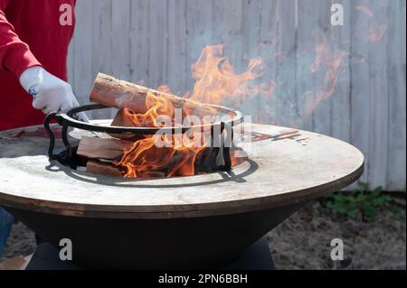 Runder Grill in Form einer Schüssel mit Feuer im Inneren. Ein Mann bereitet ein Herd für ein Barbecue vor. Brennholz brennt in der Schüssel des Herds. Stockfoto