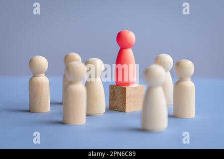 Rote weibliche Holzpuppe, die auf einem quadratischen Blockpodium steht und von anderen Puppen umgeben ist. Weibliches Führungskonzept. Stockfoto