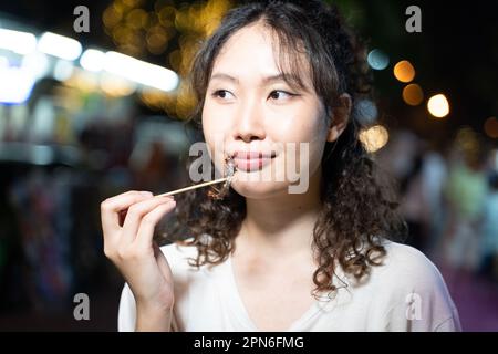 Porträt eines asiatischen Allesreisenden mit einem gebratenen Insektenstiel auf der Straße in Bangkok, Thailand. Weibliche Touristen, die Südostasien erkunden. - Kopierraum Stockfoto