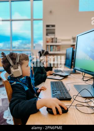 Elementare Brüder, die in der Bibliothek mit Kopfhörern auf Computern spielen Stockfoto