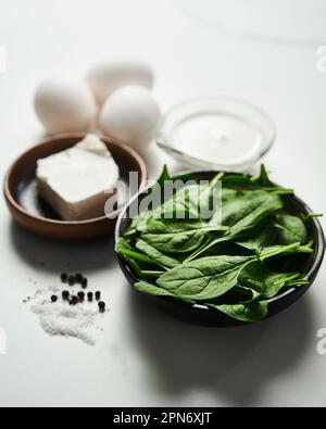 Nahaufnahme von Inhaltsstoffen für gesunden Spinat und Feta-Omelette. Eier mit schwerer Sahne, frische Baby-Spinatblätter in der schwarzen Keramikschüssel, weiß Stockfoto