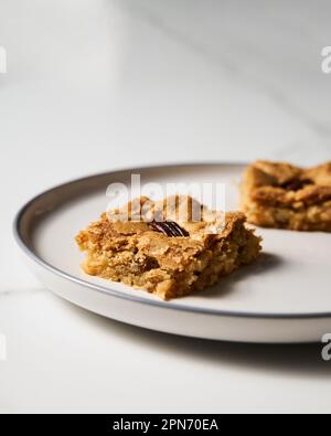 Zwei Quadrate Blondie Kekse mit Pekannüssen auf weißem Teller. Weiße Schokolade, süß und lecker, kaute Keksriegel. Schließen Stockfoto