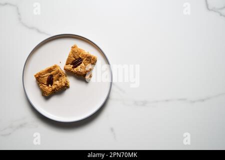 Zwei Quadrate Blondie Kekse mit Pekannüssen auf weißem Teller. Weiße Schokolade, süß und lecker, kaute Keksriegel. Draufsicht, Kopierbereich Stockfoto