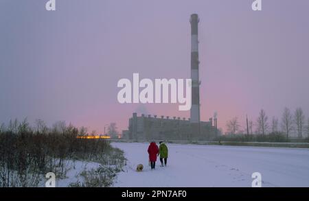 Eine Frau in Rot und eine Frau in Grün gehen mit den Hunden in einem Schneesturm vor dem Hintergrund eines Heizungsraums und gelber Lichter von Gewächshäusern Stockfoto