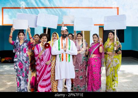 Ein indischer Politiker, der namaste mit einer Gruppe traditioneller Frauen mit Platzkarte macht. Konzept von Wahl und Abstimmung. Stockfoto