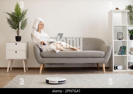 Junge lächelnde Frau im weißen Bademantel, die auf dem Sofa mit einem Laptop liegt und den Roboter-Staubsauger über die Fernbedienung läuft. Smart-Home-Gerät hält den Teppich sauber, während der Besitzer sich im Studio-Apartment ausruht. Stockfoto
