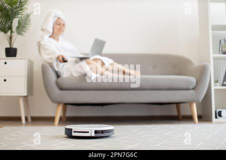 Weißer automatischer Roboter-Staubsauger, der den Teppich staubfrei hält, während sich die Besitzerin mit dem tragbaren Computer auf der gemütlichen Couch im Innenbereich entspannt. Wireless-Technologie-Gerät für eine gesunde Umgebung in luftigen Apartments. Stockfoto