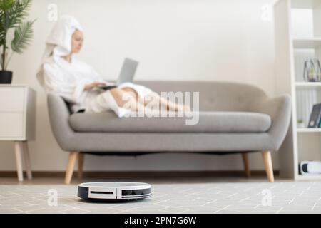 Weißer automatischer Roboter-Staubsauger, der den Teppich staubfrei hält, während sich die Besitzerin mit dem tragbaren Computer auf der gemütlichen Couch im Innenbereich entspannt. Wireless-Technologie-Gerät für eine gesunde Umgebung in luftigen Apartments. Stockfoto