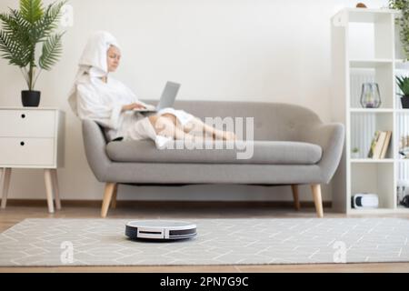 Weißer automatischer Roboter-Staubsauger, der den Teppich staubfrei hält, während sich die Besitzerin mit dem tragbaren Computer auf der gemütlichen Couch im Innenbereich entspannt. Wireless-Technologie-Gerät für eine gesunde Umgebung in luftigen Apartments. Stockfoto