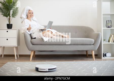 Junge lächelnde Frau im weißen Bademantel, die auf dem Sofa mit einem Laptop liegt und den Roboter-Staubsauger über der Fernbedienung läuft und den Daumen nach oben zeigt. Stockfoto