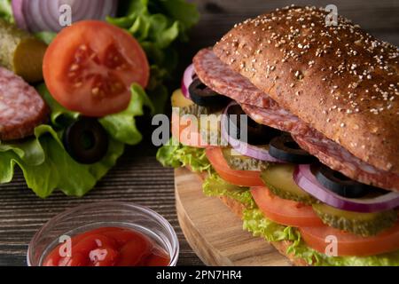 Fotozwiebeln, eingelegte Gurken, Oliven, Salami, grüner Salat auf einem Brötchen auf dem Tisch Stockfoto