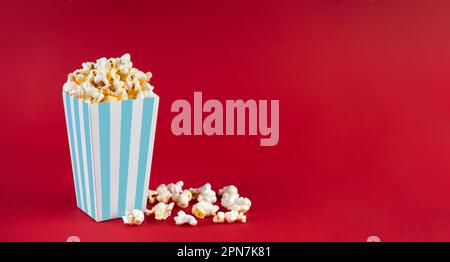 Türkisfarbener, weiß gestreifter Kartoneimer mit leckerem Käsepopcorn, isoliert auf rotem Hintergrund. Schachtel mit Streuung von Popcornkörnern. Fast Food, Filme, Stockfoto