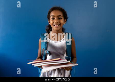 Porträt eines fröhlichen, birassischen Schulmädchens mit lächelnden Notizbüchern auf blauem Hintergrund und Kopierbereich Stockfoto
