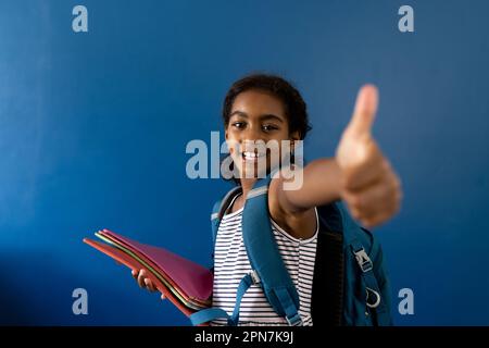 Porträt eines glücklichen, birassischen Schulmädchens, das Daumen-hoch-Zeichen mit Notizbüchern mit Kopierraum macht Stockfoto