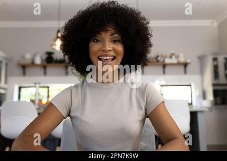 Porträt einer glücklichen, birassischen Frau mit Afro, die zu Hause sitzt, Videoanrufe macht und lächelt Stockfoto