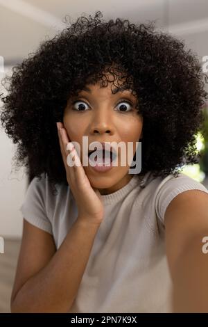 Porträt einer überraschten, birassischen Frau mit Afro zu Hause, die Videoanrufe macht und ihr Gesicht schockiert berührt Stockfoto