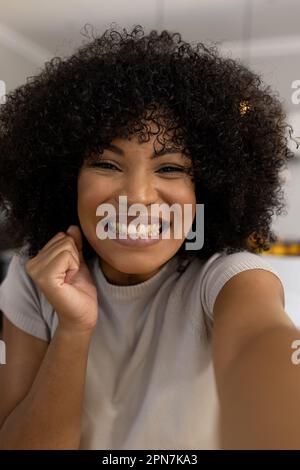 Porträt einer glücklichen, aufgeregten, birassischen Frau mit Afro zu Hause, die Videoanrufe macht und lächelt Stockfoto