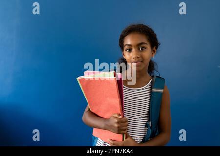 Porträt eines fröhlichen, birassischen Schulmädchens mit Notizbüchern auf blauem Hintergrund und Kopierbereich Stockfoto
