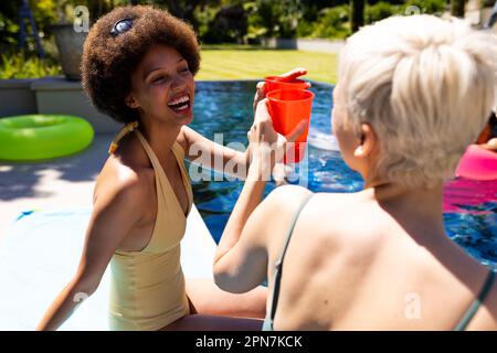 Fröhliche, vielfältige weibliche Freunde, die eine Poolparty feiern, einen Toast ausbringen und im Garten lächeln Stockfoto
