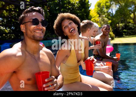 Fröhliche, vielfältige Gruppe von Freunden, die eine Poolparty veranstalteten und Plastikbecher im Garten hielten Stockfoto