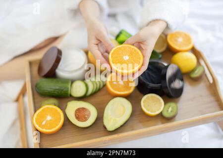 Konzentrieren Sie sich auf die Hälfte der saftigen Orangencitrus, die von den weiblichen Händen über dem Tablett mit frischem Obst und Glasgefäßen gehalten wird. Fruchtbestandteile in Kosmetika werden zum Hautpflegetrend des exklusiven Lebensstils. Stockfoto