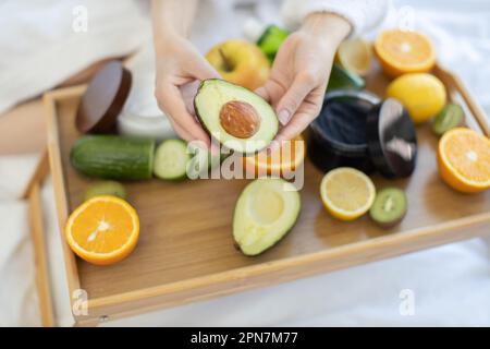 Nahaufnahme des grünen Avocados in Frauenhänden, die über geschnittenem natürlichem Obst und Gemüse gehalten werden, das auf einem Tablett unter Schönheitsprodukten platziert wird. Organische Inhaltsstoffe für gesundes Hautglühen. Stockfoto