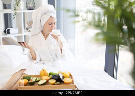 Attraktive, gesunde Frau nach dem Baden mit frischem Aroma von leichter Feuchtigkeitscreme und Sonnenlicht vom Panoramafenster des Schlafzimmers. Fruchtschichten als Hauptbestandteil der Schönheitstherapie. Stockfoto