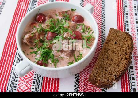 Köstlicher Rindereintopf mit Kirschen, Schwarzbrot, in einer weißen Schüssel, auf einer bestickten ukrainischen Serviette. Stockfoto