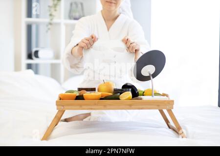 Nahaufnahme der jungen Frau im Bademantel halten Papiertuchmaske über dem Betttablett mit frischem Obst und Kosmetikprodukten im Schlafzimmer-Interieur. Kosmetische Gesichtsmasken, die mit Hautpflegeprodukten beladen werden. Stockfoto