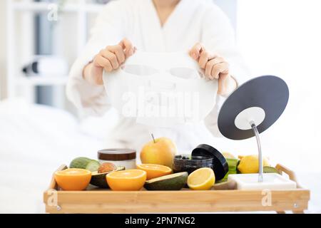 Nahaufnahme der jungen Frau im Bademantel halten Papiertuchmaske über dem Betttablett mit frischem Obst und Kosmetikprodukten im Schlafzimmer-Interieur. Kosmetische Gesichtsmasken, die mit Hautpflegeprodukten beladen werden. Stockfoto