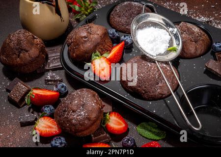 Schokoladen-Muffins mit Beeren. Muffin-Pfanne mit hausgemachten süßen, dunklen Schokoladen-Cupcakes mit Kakaosoße, Zuckerpulver und frischen Erdbeeren, Blaubeeren Stockfoto