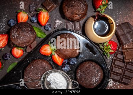 Schokoladen-Muffins mit Beeren. Muffin-Pfanne mit hausgemachten süßen, dunklen Schokoladen-Cupcakes mit Kakaosoße, Zuckerpulver und frischen Erdbeeren, Blaubeeren Stockfoto