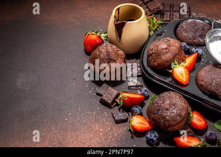 Schokoladen-Muffins mit Beeren. Muffin-Pfanne mit hausgemachten süßen, dunklen Schokoladen-Cupcakes mit Kakaosoße, Zuckerpulver und frischen Erdbeeren, Blaubeeren Stockfoto